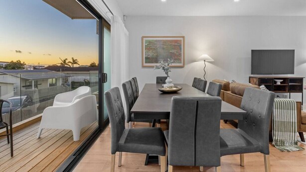 A modern living room with a dining area, large windows, a flat-screen TV, and a balcony with a view of the sunset. - ﻿Premium Sunset Townhouse | 6 Aircons near Airport (Auckland South)