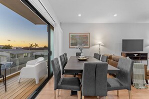 Un salon moderne comprenant un coin repas, de grandes fenêtres, un téléviseur à écran plat et un balcon offrant une vue sur le coucher de soleil.