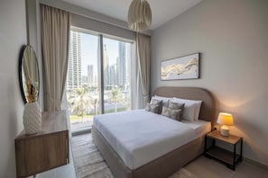 A modern hotel room with a large bed, a bedside table with a lamp, a wall-mounted mirror, and a view of the cityscape through large windows.