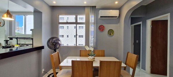 Un comedor con una mesa y sillas, un equipo de aire acondicionado, una ventana con vistas a edificios y un ventilador.