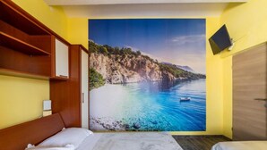 A hotel room with a large scenic mural of a coastal landscape, a wooden headboard, a bed with white bedding, and a small television mounted on the wall. - Apartments Ribarević (Makarska)
