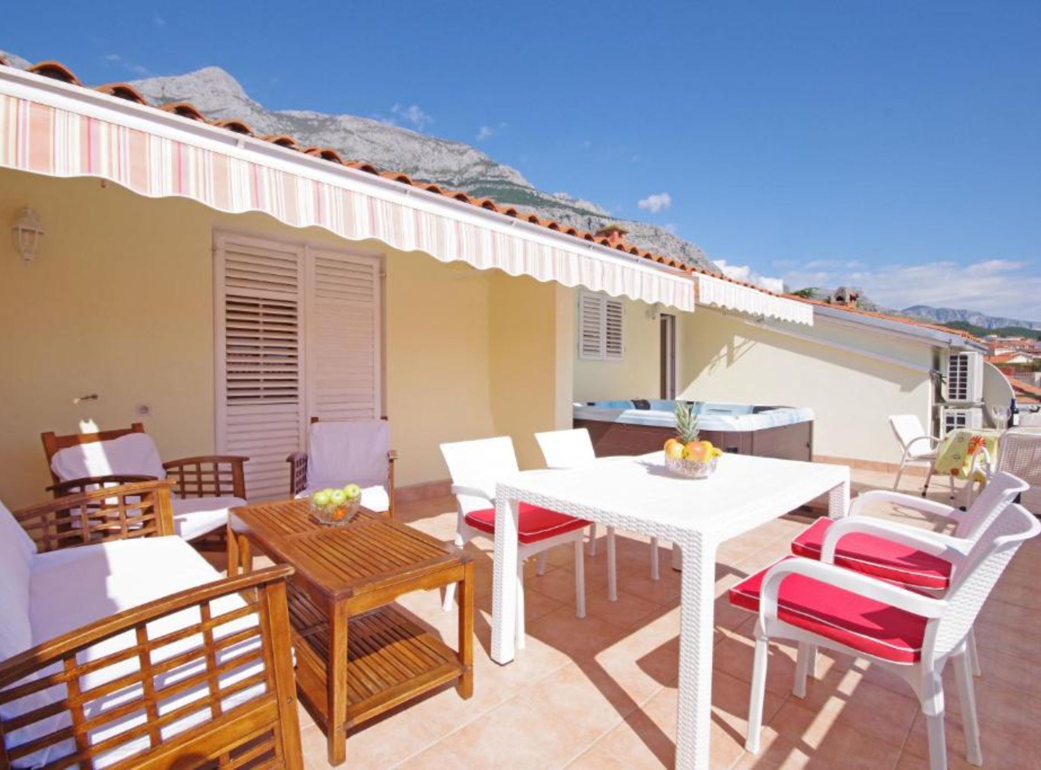 A terrace with wooden furniture and white chairs, a table with fruit, and a view of mountains.