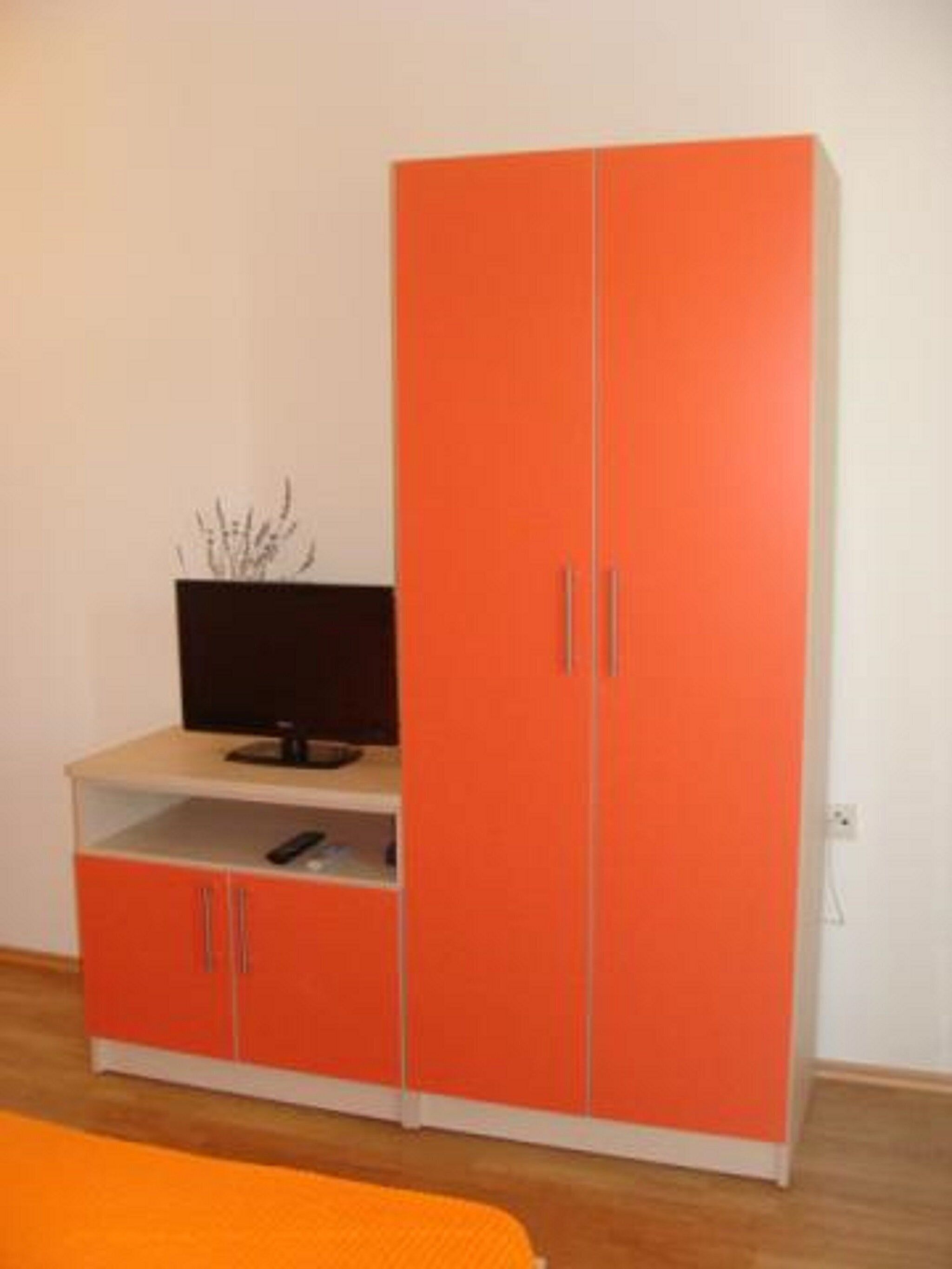 Ein Zimmer mit einem großen, orangefarbenen Kleiderschrank, einem Flachbildfernseher auf einer niedrigen Ablage und einer kleinen Pflanze auf dem Boden.