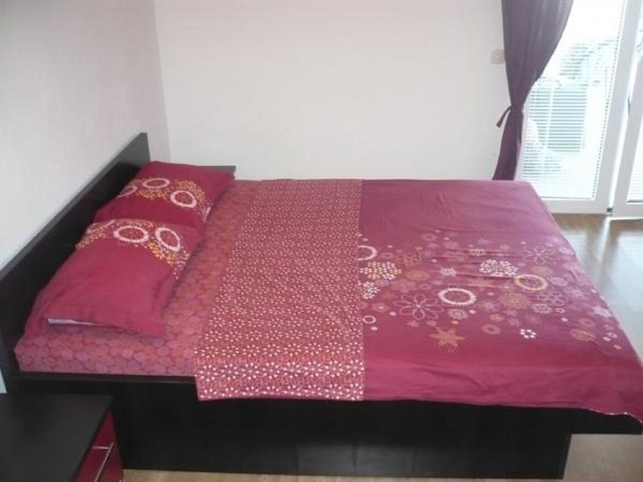 A bed with a patterned bedspread and pillows, a black headboard, and a window with blinds.