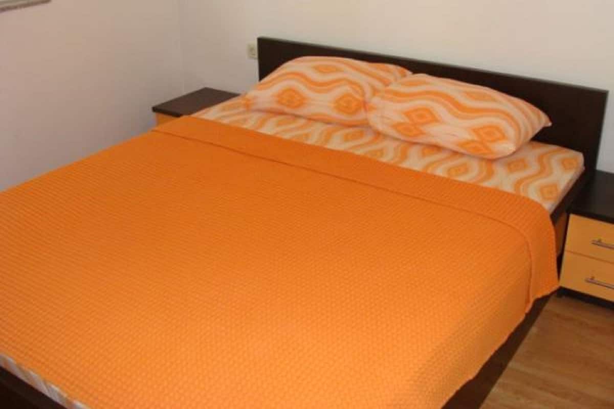 A bed with an orange blanket and patterned pillows, a wooden nightstand, and a drawer unit.