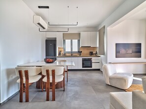 A modern kitchen with white cabinets, a built-in oven, and a dining area with a white table and chairs. - Parousia Luxury Apartments (Paros)