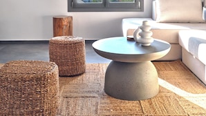 A modern living room with a round table, two woven poufs, a sofa, and a large window offering a view of a lake and hills. - Parousia Luxury Apartments (Paros)