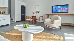 A modern living room with a white coffee table, a TV mounted on the wall, and a kitchen area with white cabinets and a black refrigerator. - Parousia Luxury Apartments (Paros)