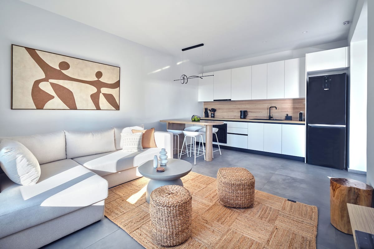 A modern living room with a sofa, coffee table, and ottomans, a kitchen with white cabinets and a black refrigerator, and a large abstract painting on the wall.