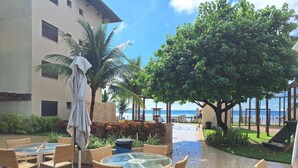 A patio with round tables and wicker chairs, overlooking a pool and a view of the ocean. - 2 bedroom apartment in Ipojuca - PE (Ipojuca - PE)