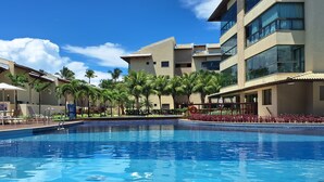 A swimming pool with tiled edges, palm trees, and a multi-story building with balconies. - 2 bedroom apartment in Ipojuca - PE (Ipojuca - PE)