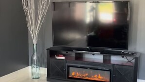 Un salon moderne avec un téléviseur à écran plat fixé à un meuble noir, une cheminée située sous le téléviseur, un tapis noir à poils longs et un coussin rouge sur un canapé en cuir.