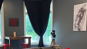 Un salon moderne avec un canapé en cuir noir, des coussins rouges, un tapis foncé, un trépied et une pancarte « MANGUEZ-MOI » accrochée au mur.