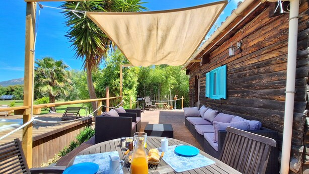 Un espace extérieur aménagé avec des meubles en bois, une table dressée avec des assiettes, une carafe de jus d’orange et une bouteille d’eau.