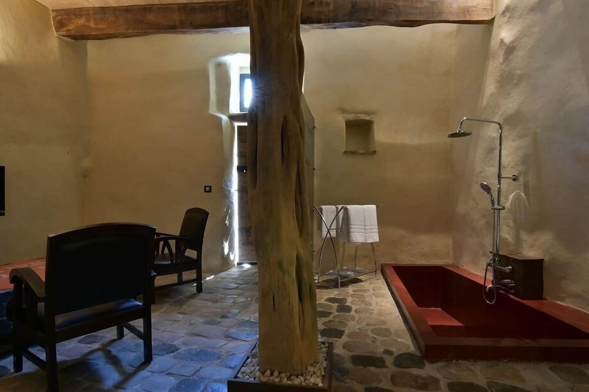 A rustic bathroom with a stone floor, a wooden tree trunk centerpiece, a red bathtub, and a shower area.
