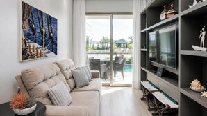 A modern living room with a sofa, a flat-screen TV, a bookshelf, and a view of an outdoor pool area. - 350m from Jurerê Beach | Swimming Pool #JA36 (Florianópolis)