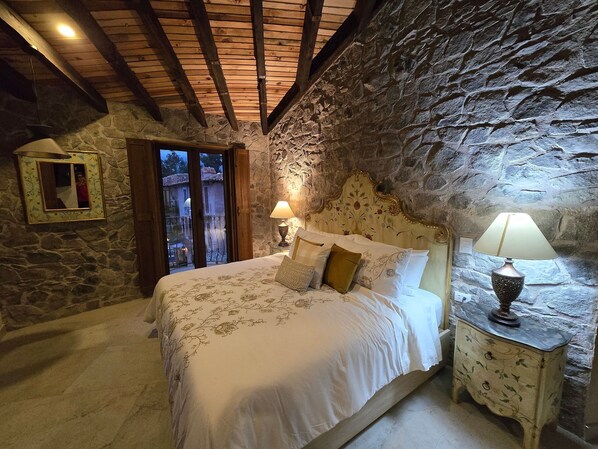 A bedroom with a stone wall, a wooden ceiling, a bed with floral bedding, and a nightstand with a lamp. - Loft Michelangelo (Val'Quirico)
