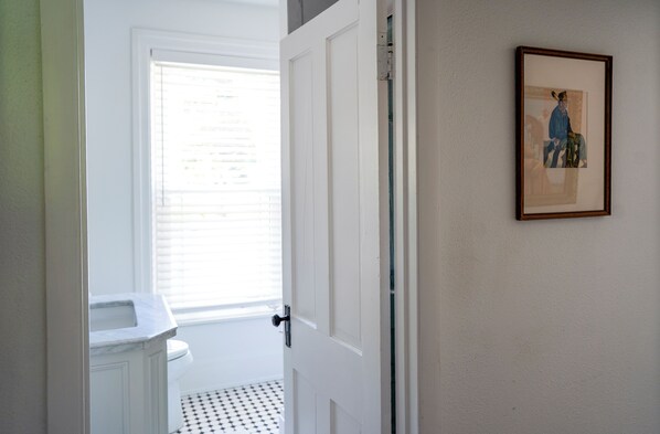 Un baño con lavamanos y retrete blancos, una puerta que da a otra habitación y un cuadro en la pared.