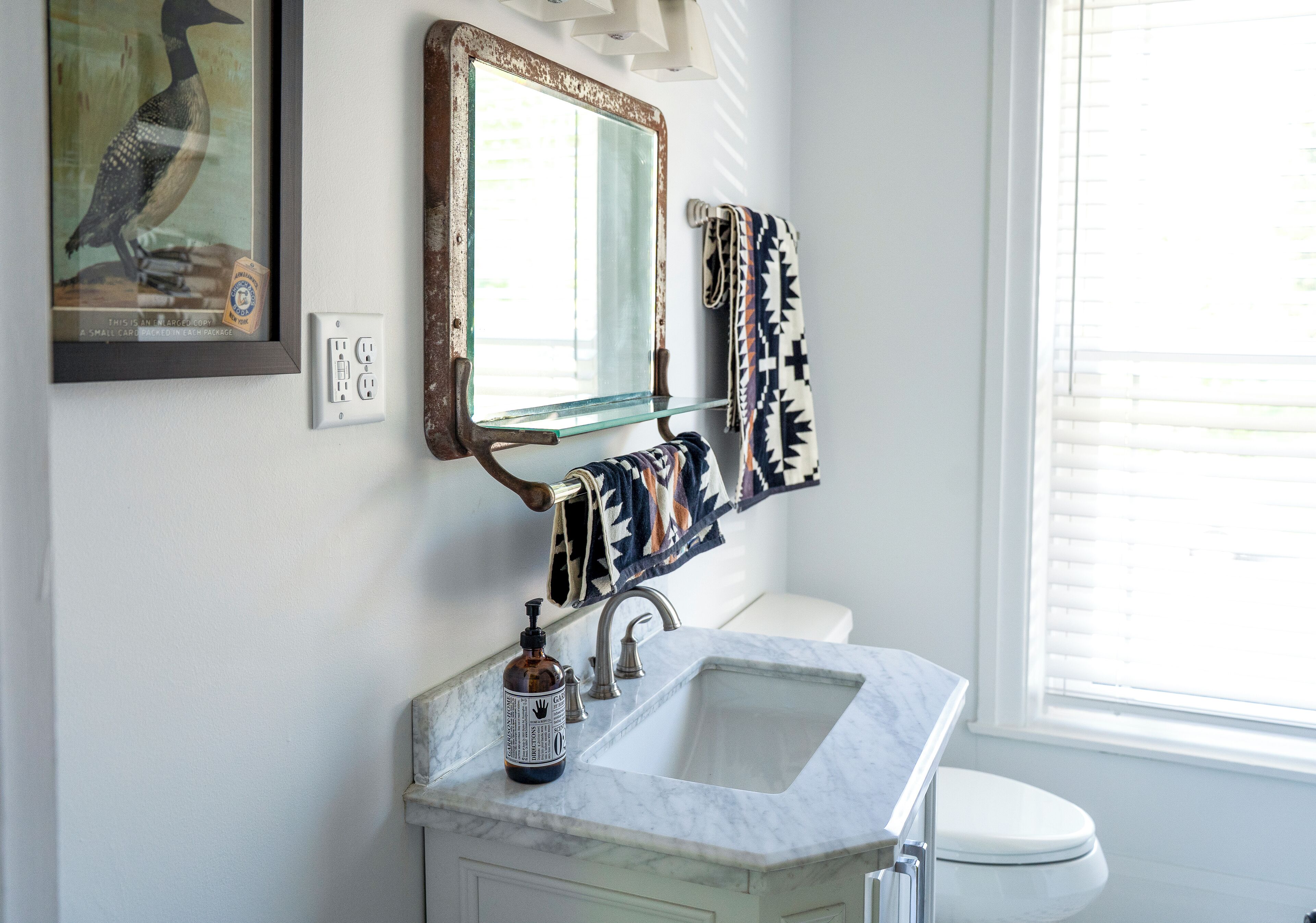 Une salle de bain avec un lavabo en marbre, une étagère en verre, un miroir doté d’un cadre rustique et un porte-serviettes avec des serviettes à motifs.