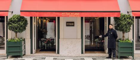 Entrada de hotel com toldo vermelho, um letreiro indicando "Mythic & Sana Downtown Suites" e um porteiro à porta.
