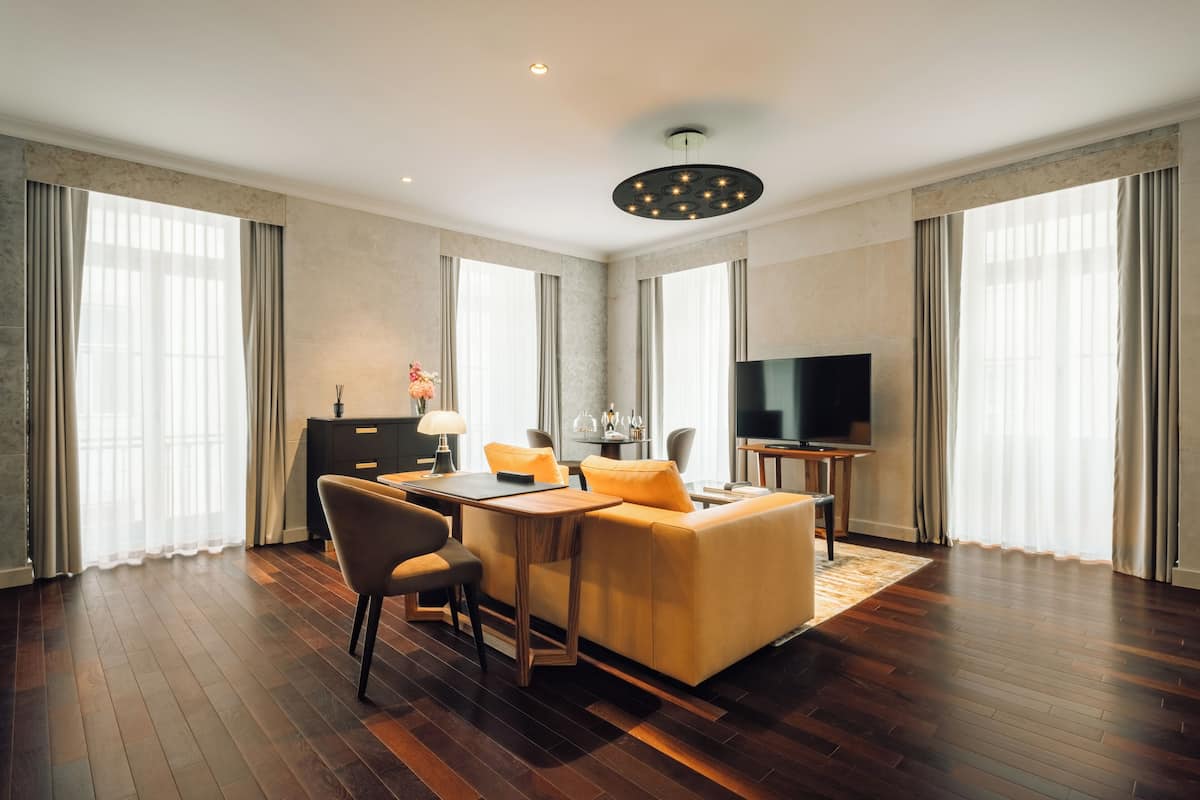 Una habitación de hotel moderna con piso de madera, un sofá, una mesa de comedor con sillas, un televisor y ventanas grandes con cortinas.
