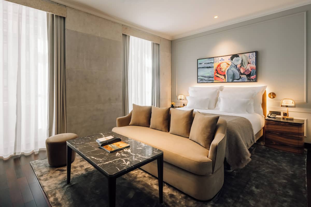 Una habitación de hotel moderna con sofá, una mesita, cama con ropa blanca, mesita de noche y un cuadro en la pared.