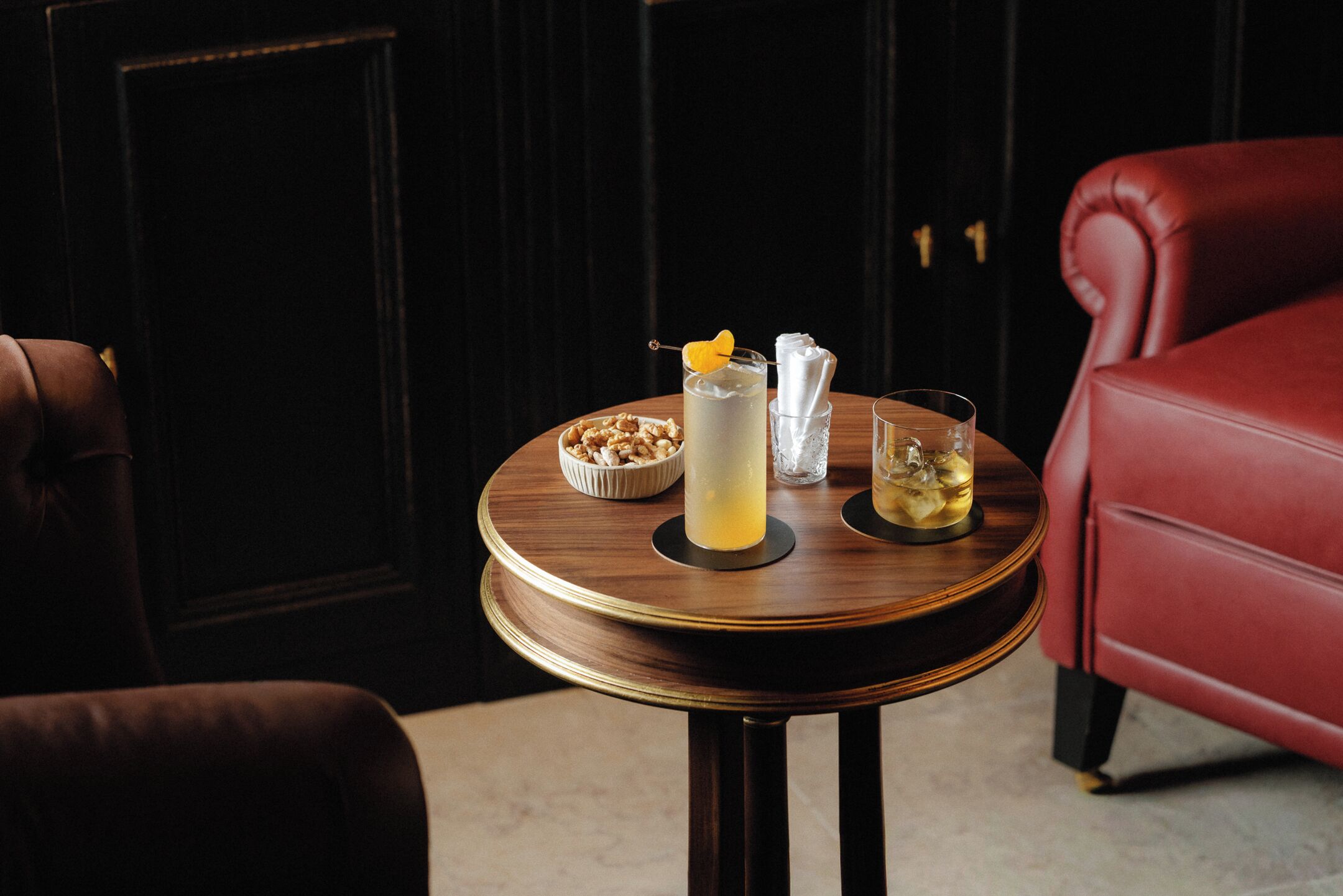 A round wooden table with a drink, a bowl of nuts, and a glass of water in a room with dark wood paneling and red leather chairs.