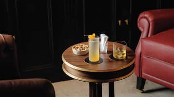 Una mesa redonda de madera con una bebida, un cuenco de nueces y un vaso de agua en una habitación con paneles de madera oscura y sillas de cuero rojo.