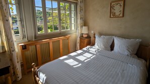 A bedroom with a large window, a wooden headboard, a bedside table with a lamp, and a framed picture on the wall. - Auberge des Chasseurs (Echenevex)
