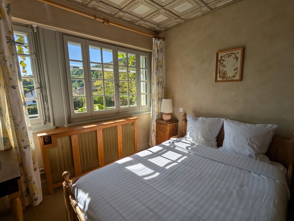 A bedroom with a large window, a wooden headboard, a bedside table with a lamp, and a framed picture on the wall. - Auberge des Chasseurs (Echenevex)