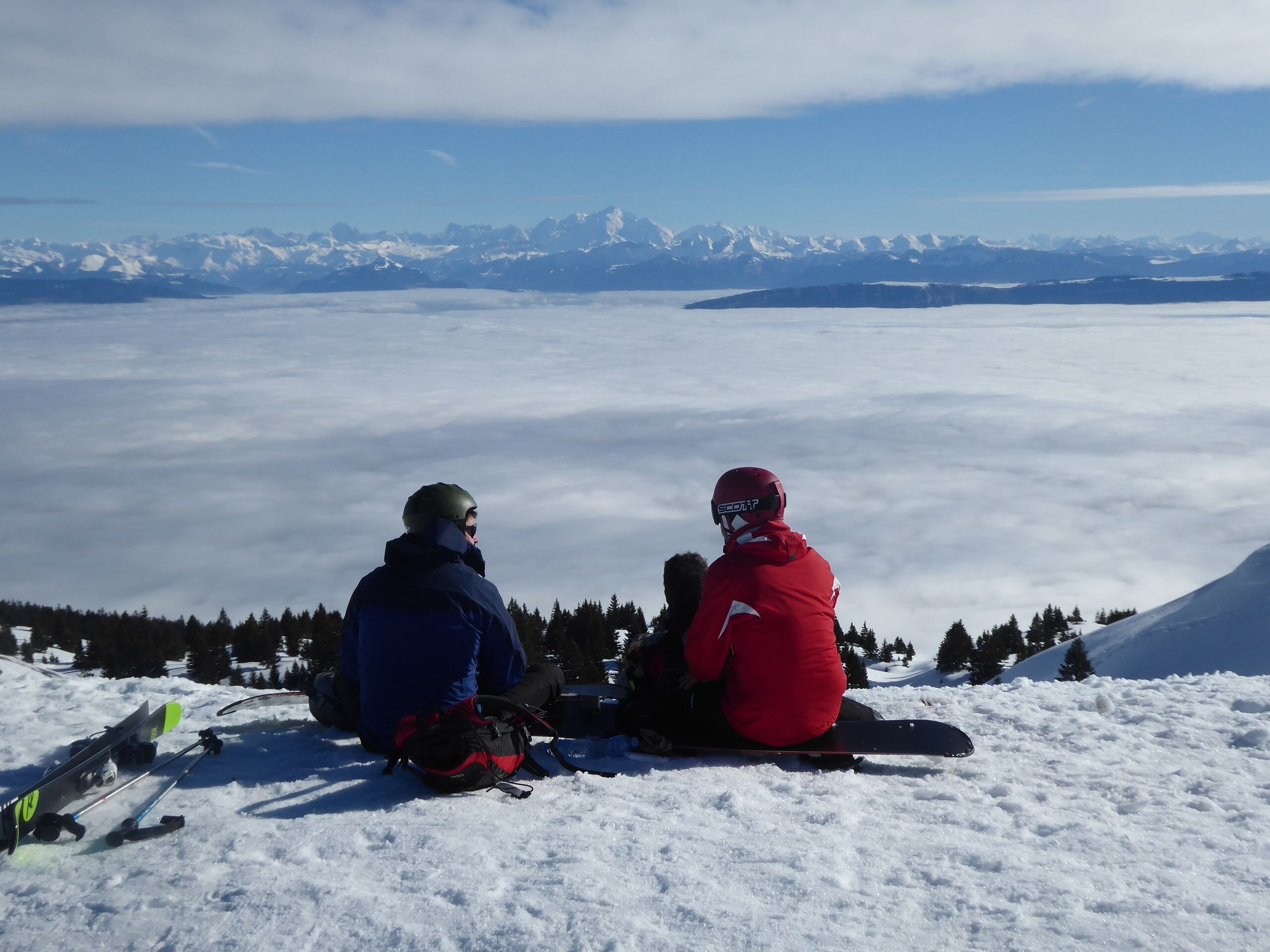 Two people sitting on a snowy mountain with skis and a snowboard nearby, overlooking a vast snowy landscape and distant mountains.