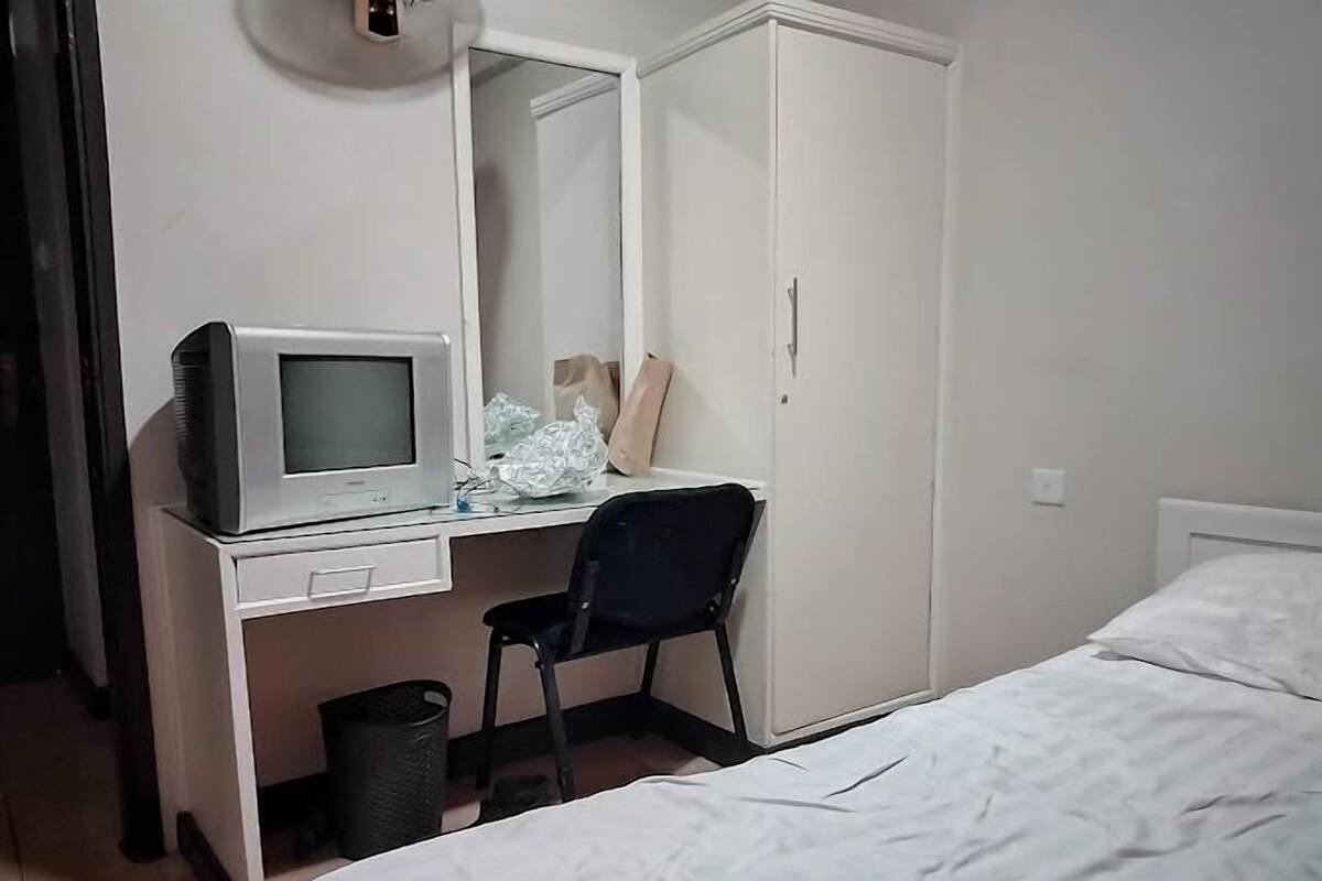 A bedroom with a bed, a desk with a computer, a chair, a wardrobe, and a trash bin.