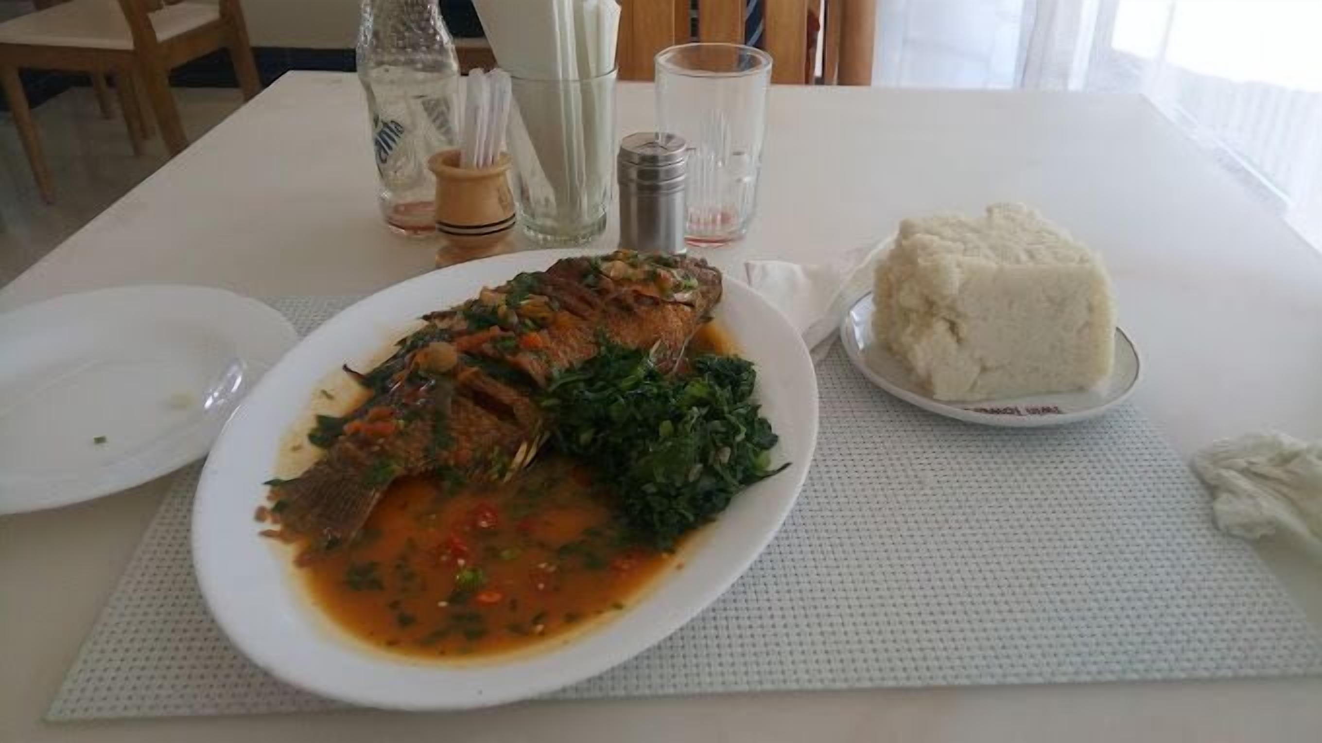 A dining table with a plate of fish, greens, and a mound of white food, possibly rice, with condiments and glasses in the background.