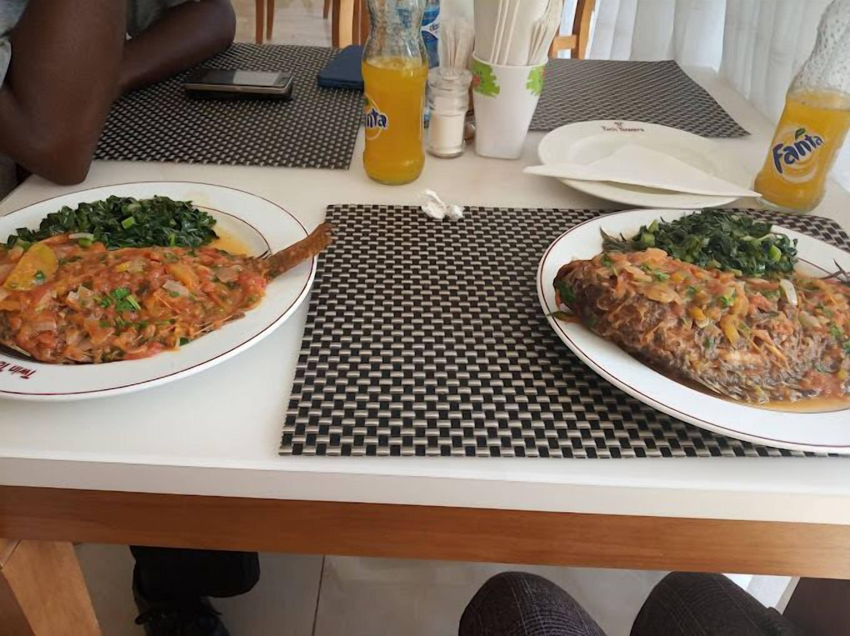 A dining table with two plates of fish and greens, a bottle of Fanta, and a mobile phone on the table.