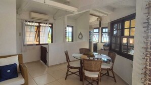 A dining area with a round table and four chairs, a wicker sofa with cushions, and a glass cabinet with dishes. - Suítes Azul Marinho (Ilhabela)