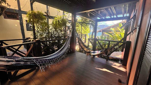 A hammock on a wooden deck with a view of lush greenery and a building in the background. - Suítes Azul Marinho (Ilhabela)