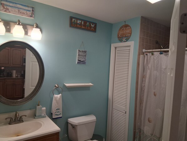 A bathroom with a light blue wall, a large mirror, a sink with a faucet, a toilet, a shower curtain, and various wall decorations including a 'RELAX' sign and a 'This Home' sign. - Reeds beach retreat 2 bedroom with water view (Reeds Beach)