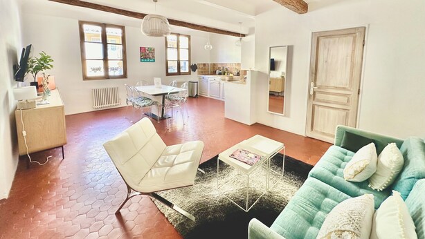 A spacious living room with a white sofa, a green sofa, and a white chair. There is a dining table with chairs, a kitchen area, and a large window.
