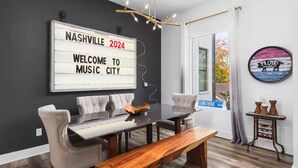 A modern dining area with a glass table, cushioned chairs, and a wooden bench. A large sign on the wall reads "NASHVILLE 2024 Welcome to Music City."