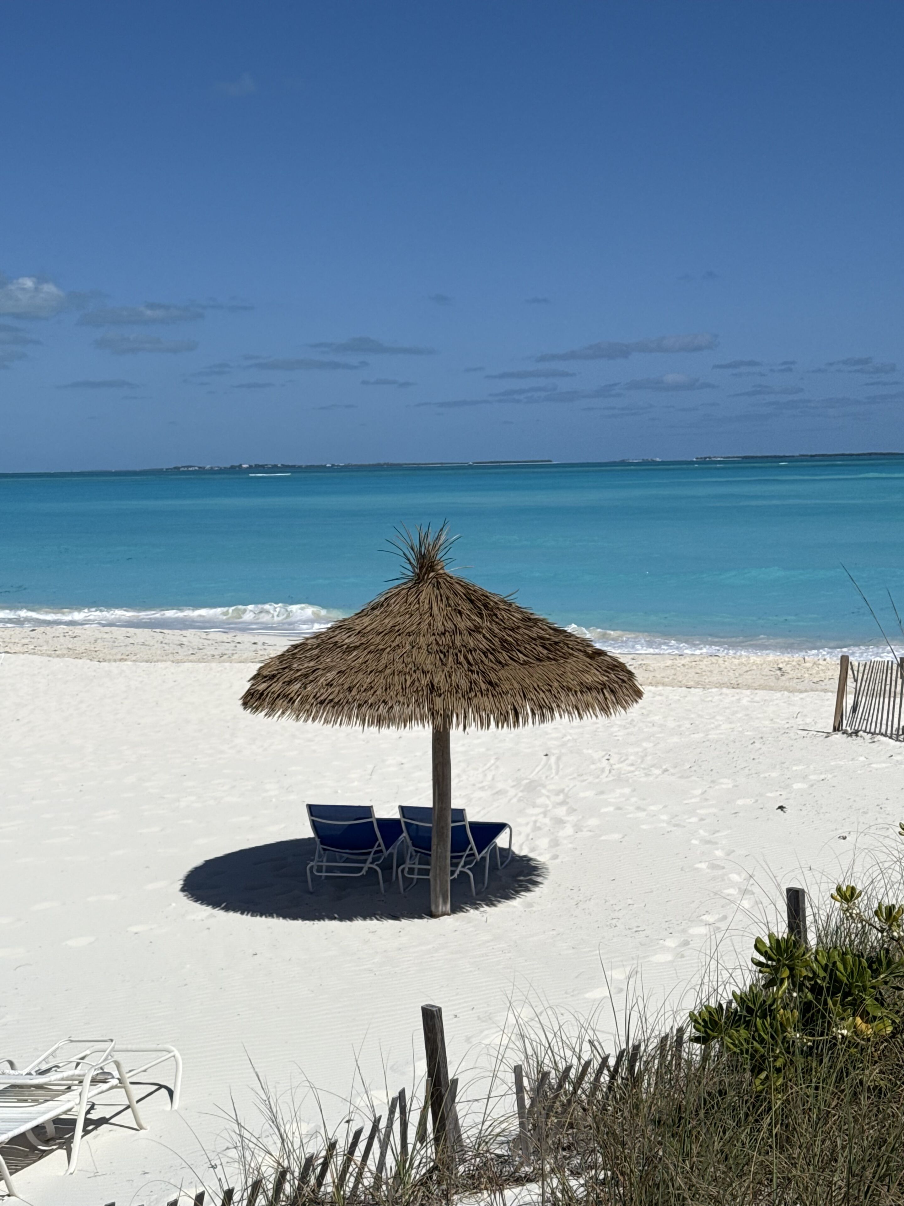A beach with a thatched umbrella, two blue lounge chairs, and a wooden fence.