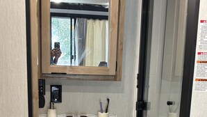 A bathroom with a white sink, wooden cabinets, a mirror, and a glass shower enclosure. - Timberwolf in the Park (Pensacola)