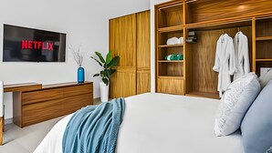 Ein modernes Schlafzimmer mit einem großen Bett, einem Holzschrank, einem Schreibtisch mit einem Fernseher, der 'NETFLIX' anzeigt, und einer Topfpflanze.