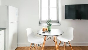 Una cocina moderna con un refrigerador blanco, una mesa de comedor con dos sillas blancas, una planta en maceta y un televisor de pantalla plana montado en la pared.
