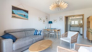 A living room with a grey sofa, a wooden coffee table, wicker chairs, a dining table with chairs, and a kitchen visible through an open door. - Beau Rivage - 7min à pied de la Plage (Carqueiranne)