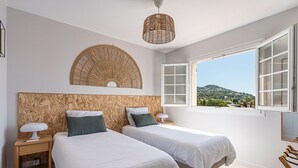 A bedroom with two beds, a large woven headboard, bedside lamps, and a window with a view of a hilly landscape. - Beau Rivage - 7min à pied de la Plage (Carqueiranne)