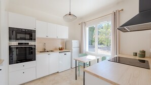 A modern kitchen with white cabinets, a built-in oven, a dishwasher, and a dining area with a table and chairs. - Lou Vaou - Maison Climatisée avec Piscine (Carcès)