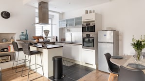A modern kitchen with white cabinets, a central island with bar stools, a built-in oven, and a refrigerator. - Le Repos des Vagues - 4min de la Plage (Dinard)