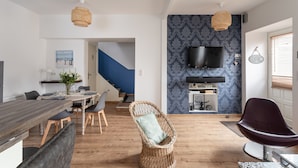A modern living space with a dining area, a wicker chair, a flat-screen TV, and a staircase. - Le Repos des Vagues - 4min de la Plage (Dinard)
