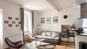 A modern living room with a grey sofa, wicker chair, and a small table. There are framed pictures on the wall and a kitchen area with bar stools. - Le Repos des Vagues - 4min de la Plage (Dinard)