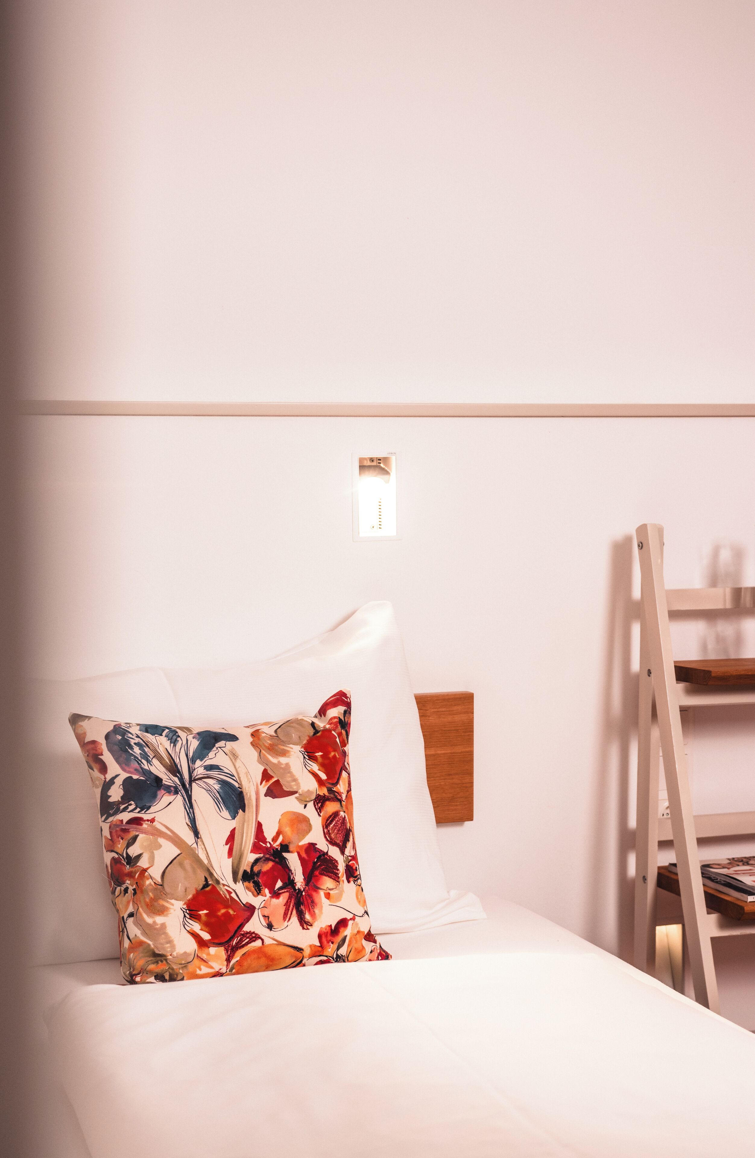 A neatly made bed with a floral pillow, a wooden headboard, and a white wall with a small light fixture.